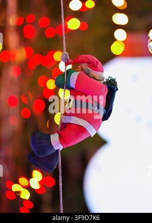 un'immagine umoristica di Babbo Natale appeso a una corda in un ambientazione festiva all'aperto, circondato da tessuti colorati e luci bokeh, creando un'atmosfera vivace Foto Stock