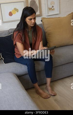 Donna adulta indiana seduta su un divano grigio in salotto con un tablet, indossa una maglietta olivastra, jeans Foto Stock