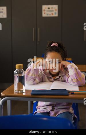 Bambino afroamericano in camicia a quadri che appoggia la testa sulle mani a un banco di classe con quaderno e bottiglia Foto Stock