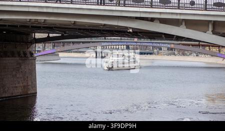Mosca, Russia – 13 marzo 2022: Barca che naviga sotto un ponte su un fiume con lo skyline della città sullo sfondo, architettura urbana visibile Foto Stock