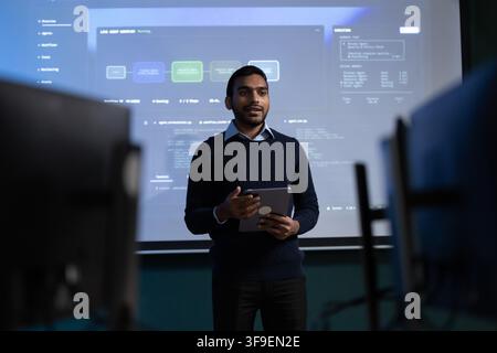 Giovane professionista indiano tiene un tablet mentre presenta un flusso di lavoro software a colleghi in una sala riunioni, con schermi di computer e ascoltatori Foto Stock