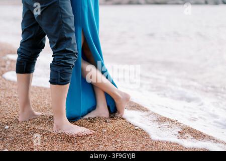 Una vista ravvicinata di una coppia in piedi a piedi nudi su una spiaggia di ciottoli, con onde gentili che lambiscono i loro piedi. La donna indossa un vestito blu con uno spacco, e l'uomo ha i pantaloni arrotolati. La scena cattura un momento romantico e intimo in riva al mare. Foto Stock