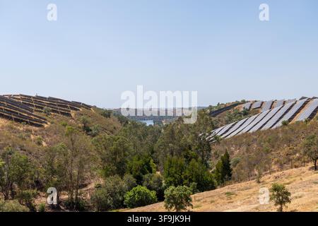 Vasti parchi solari sono installati sui pendii di un terreno asciutto e collinoso con alberi sparsi e uno specchio d'acqua in lontananza Foto Stock