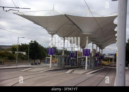 Tallaght, Contea di Dublino, Irlanda, 15 agosto 2026 – La distintiva tenda a tetto bianca, le banchine a isola, le linee elettriche aeree, i segnali di direzione e i validatori dei biglietti alla fermata Belgard Luas della linea rossa di Dublino. Belgard funge da snodo per i servizi di tram verso Tallaght e Saggart, con servizi verso il centro città di Dublino nella direzione opposta. La fermata è stata riconfigurata per il ramo Saggart con una linea aggiuntiva, banchine modificate e una tettoia che copre le banchine a isola, rafforzando il suo ruolo di interscambio della metropolitana leggera nel sud-ovest di Dublino. Foto Stock
