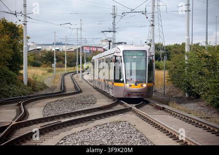 Tallaght, Contea di Dublino, Irlanda, 15 agosto 2026 – La distintiva tenda a tetto bianca, le banchine a isola, le linee elettriche aeree, i segnali di direzione e i validatori dei biglietti alla fermata Belgard Luas della linea rossa di Dublino. Belgard funge da snodo per i servizi di tram verso Tallaght e Saggart, con servizi verso il centro città di Dublino nella direzione opposta. La fermata è stata riconfigurata per il ramo Saggart con una linea aggiuntiva, banchine modificate e una tettoia che copre le banchine a isola, rafforzando il suo ruolo di interscambio della metropolitana leggera nel sud-ovest di Dublino. Foto Stock