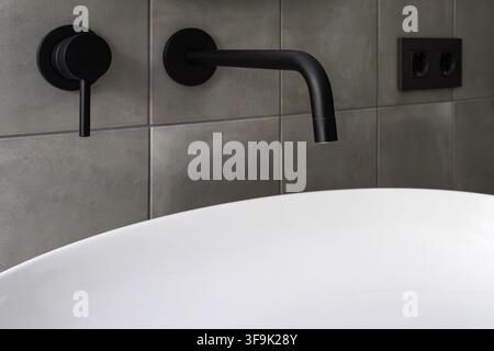 Interno di bagno minimalista con rubinetto della vasca montato a parete nero opaco, vasca bianca e piastrelle grigio caldo Foto Stock