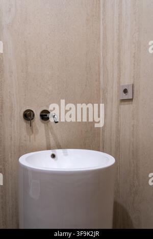 Lavandino rotondo minimalista con rubinetto a parete cromato contro parete in travertino beige, nessuna persona, bagno moderno Foto Stock