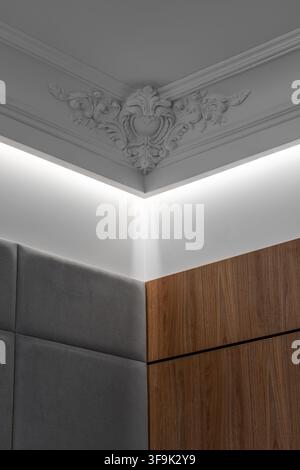 Interno moderno ed elegante con modanature classiche ornate sul soffitto, illuminazione LED indiretta calda, mobili in legno di noce e pannelli murali imbottiti Foto Stock