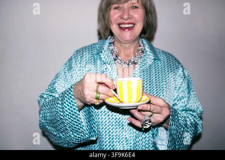 Donna matura con capelli grigi in una camicetta turchese tiene una tazza da tè a strisce gialle e bianche con piattino, sorridendo davanti a uno sfondo semplice Foto Stock