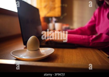 Uovo esposto in un supporto bianco su una scrivania di legno accanto a un laptop, con una persona in camicia rosa che scrive e una luce soffusa che entra da una finestra Foto Stock