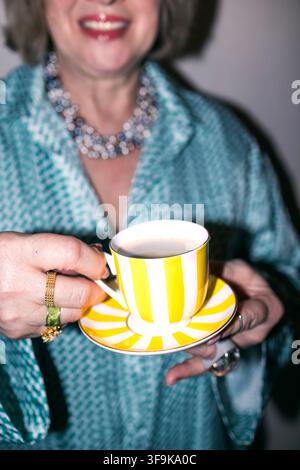 Donna con una collana statement tiene una tazza da tè e un piattino a righe gialle e bianche, indossa una camicetta a motivi turchesi in un ambiente interno illuminato con delicatezza Foto Stock