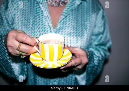 Donna tiene una tazza da tè a righe gialle e bianche con piattino, vestita con una camicetta a motivi turchesi con gioielli d'argento, su uno sfondo semplice Foto Stock