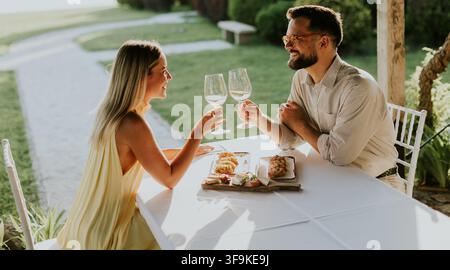 Due persone festeggiano con bicchieri alzati in un bellissimo ambiente all'aperto, condividendo un pasto delizioso e risate durante l'ora dorata. Foto Stock