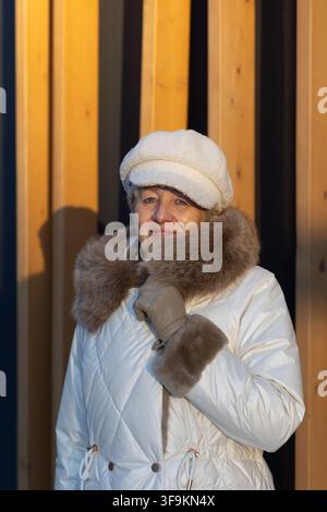Bella donna matura con eleganza senza età che sorride alla telecamera davanti a un edificio moderno in legno in una giornata invernale soleggiata. Foto Stock