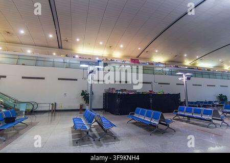 Un ampio interno del terminal aeroportuale presenta file di sedili blu per i passeggeri, una scala mobile, ringhiere in vetro, livelli commerciali e un soffitto illuminato a motivi. Foto Stock