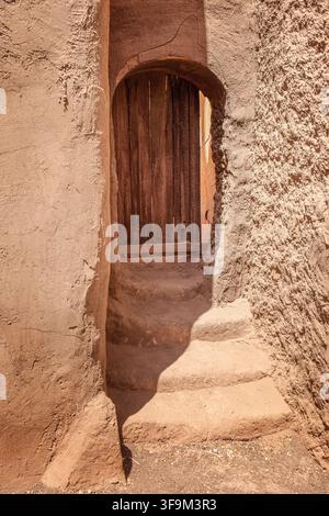 stretto passaggio di terra con gradini che portano a una porta di legno rustica in un antico edificio in mattoni di fango berbero in Marocco, verticale Foto Stock