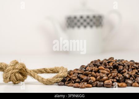 Primo piano ad angolo basso di chicchi di caffè tostati accanto a un nodo spesso di iuta, con una caffettiera di ceramica a motivi sfocata dolcemente in sottofondo e abbondante co Foto Stock