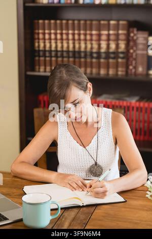 Giovane donna con occhiali scrive in un planner aperto su un tavolo di legno con un laptop e una tazza, concentrata sullo studio e l'organizzazione del suo lavoro a casa. Foto Stock