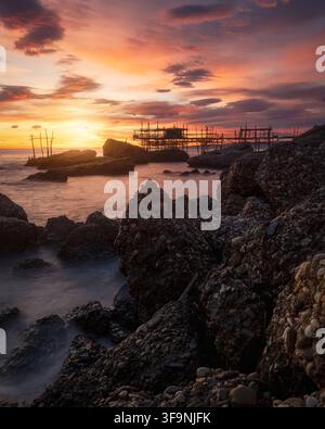 Vista della struttura di pesca storica Trabocco Spezzacatena silouettata contro un tramonto vivace sulla costa rocciosa di Rocca San Giovanni, Prov Foto Stock