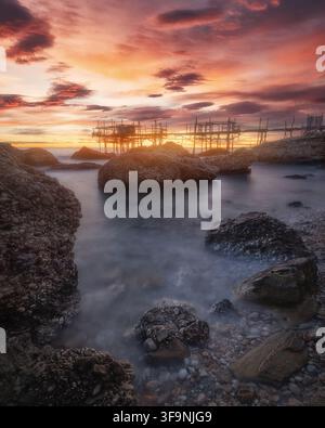 Vista del Trabocco Spezzacatena al tramonto con mare calmo, grandi rocce in primo piano e un cielo vivace arancione e viola a Rocca San Giovanni, Provi Foto Stock