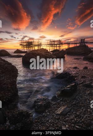 Vista del Trabocco Spezzacatena al tramonto con drammatiche nuvole arancioni sopra il mare Adriatico, costa rocciosa e onde calme a Rocca San Giovanni, Prov Foto Stock