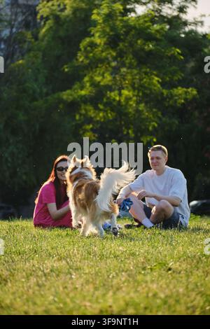 Cane che cammina sull'erba verso una coppia seduta nel parco al tramonto. Concetto di stile di vita all'aperto e animali domestici nella natura. Foto Stock