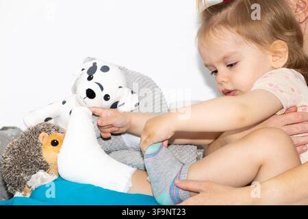 Bambina triste con la gamba rotta in una fasciatura di gesso bianca seduta sul letto con la madre e giocattoli di peluche intorno spazio copia Foto Stock
