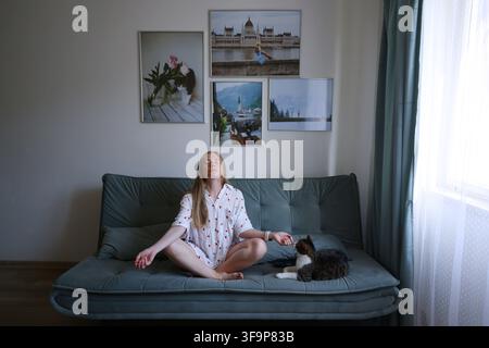 Giovane donna che si allunga dopo il risveglio seduta sul divano, routine mattutina sana e concetto di benessere. Foto Stock