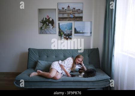 Giovane donna che si rilassa sul divano con il suo gatto e controlla lo smartphone a casa durante un momento di quieto tempo libero. Foto Stock