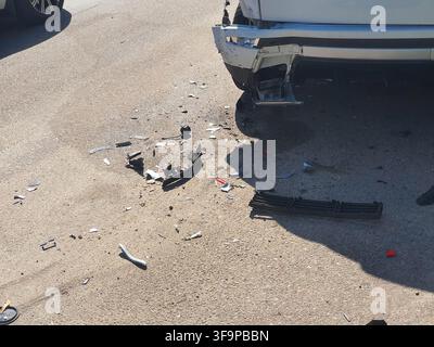 Le conseguenze di una collisione tra veicoli che mostra un SUV bianco danneggiato con un paraurti anteriore rotto e detriti sparsi sul manto stradale asfaltato. La parte anteriore destra Foto Stock