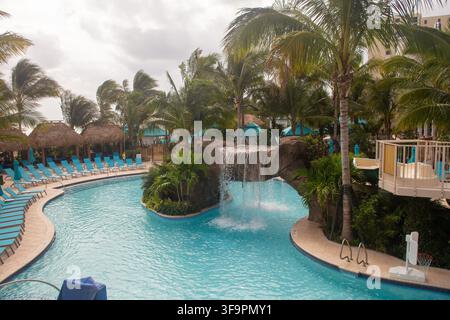 Una grande piscina in stile resort vuota con un arco a cascata in pietra artificiale, file di sdraio blu, palapa in paglia e vegetazione rigogliosa Foto Stock