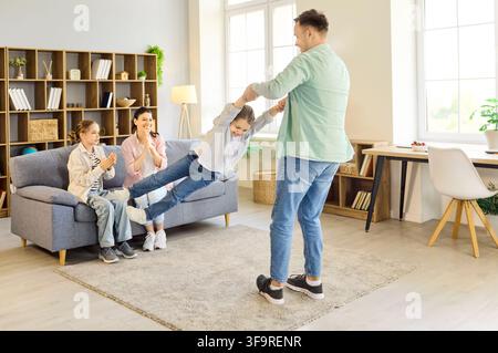 Padre che fa girare la figlia mentre la famiglia applaude in un luminoso e accogliente soggiorno a casa Foto Stock