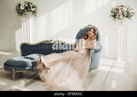 Donna incinta elegante con un lungo vestito in polvere e capelli ricci. Bella futura madre in piedi e in posa in uno studio bianco. Donna sorridente Foto Stock