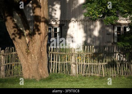 Un'affascinante fattoria in legno rustica con intricati dettagli intagliati, una tradizionale recinzione a graticcio e un grande albero in primo piano sotto una luce morbida e screziata Foto Stock