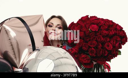 Ritratto in studio di grazioso allegro bruna giovane signora con grandi costosi bouquet di rose rosse e il mazzetto di luminose palloncini d'aria. Isolare di bianco. Smi Foto Stock