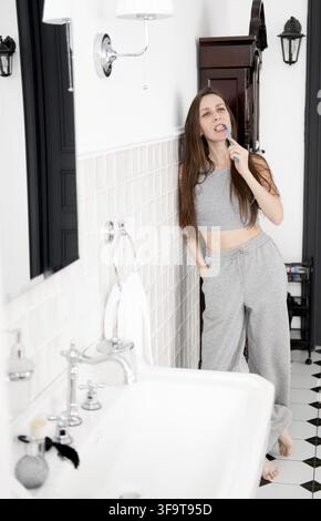 Una bella e giovane donna in pigiama grigio con apparecchio dentale è in bagno, appoggiata al muro e si lava i denti Foto Stock