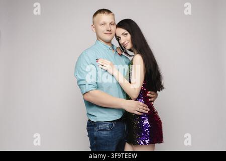 Bella giovane coppia che si abbraccia in studio. Donna attraente in abito luminoso e uomo in camicia blu e jeans che guardano la telecamera, sorridendo e p Foto Stock