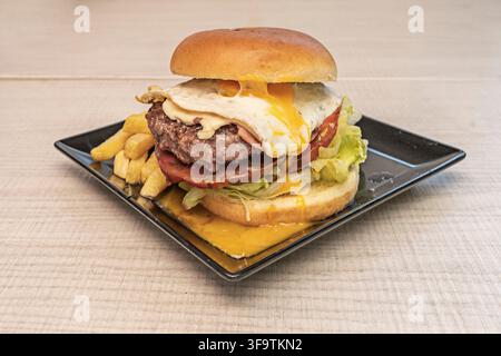 Hamburger di manzo con uovo fritto, formaggio fuso, pomodoro e lattuga servito con patatine fritte su un piatto nero Foto Stock