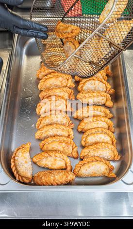 Chef che posa empanadas dorati fritti in un vassoio di metallo con un cestino di filo metallico in un ambiente da cucina. Foto Stock