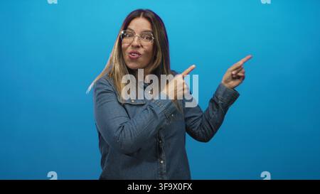 Donna di mezza età indica con entrambe le dita verso destra con bocca aperta e occhiali, indossa una camicia di jeans in uno studio blu; reazione di sorpresa. Foto Stock