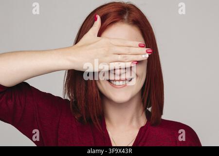 Piuttosto giovane donna con i capelli rossi nel bordo camicetta tenendo le sopracciglia e invio di bacio. Close up di bella donna con unghie rosse azienda hadns togethe Foto Stock