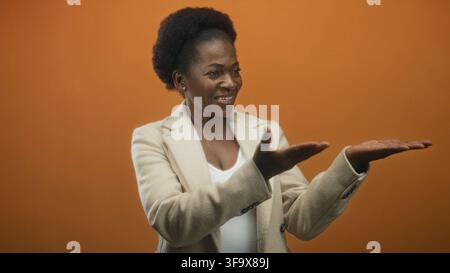 Donna che presenta palme aperte in studio con sfondo arancione e blazer beige; comunicazione di coinvolgimento. Foto Stock