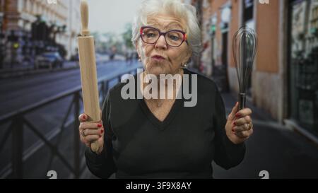Una donna anziana regge un mattarello di legno e una frusta in metallo, le mani sollevate al livello del petto in una strada trafficata della città, indossando occhiali colorati e una collana d'oro Foto Stock