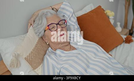 Donna anziana con occhiali colorati che dorme sul letto con la mano dietro la testa e supporto di cuscino in edificio; recupero nella serenità. Foto Stock
