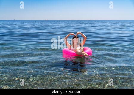 Nuoto in vacanza estiva, giovane donna che galleggia su un galleggiante rosa brillante formando un segno a cuore nell'acqua blu chiara dell'oceano. Foto Stock