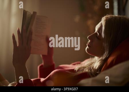 Giovane donna caucasica sdraiata e che legge un libro, concentrata sulla cura di sé e sul benessere mentale. Immagine adatta per promuovere l'alfabetizzazione, le attività ricreative e le campagne sulla salute mentale Foto Stock