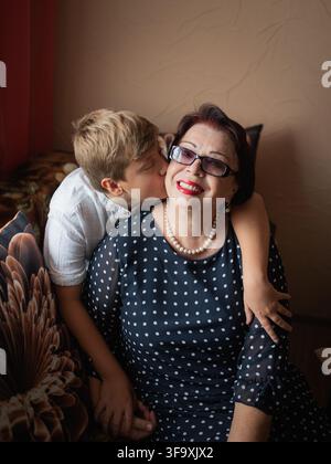 Momento commovente di amore familiare tra diverse generazioni che mostra cura e legame forte. Nonna con occhiali e vestito elegante a pois che gode Foto Stock