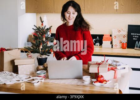 Donna in maglione di lana lavora su un laptop al tavolo della cucina accanto a un albero decorato e regali incartati. Bilanciare il lavoro durante la stagione natalizia, il tempo delle feste Foto Stock