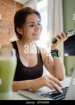Donna sorridente lavora su laptop mentre parla al telefono nell'ufficio mattutino Foto Stock