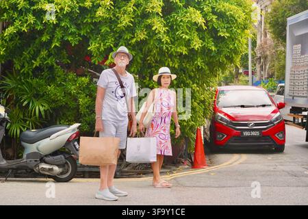 Coppia di anziani con cappelli e borse della spesa in piedi su una strada urbana a George Town Penang, Malesia Foto Stock
